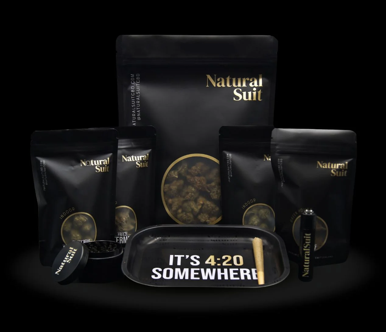 Natural-Suit-Codigo-Promocional Pack de productos de CBD de la marca Natural Suit, con bolsas de cogollos, grinder, mechero, bandeja para liar con el mensaje 'It’s 4:20 somewhere' y un porro preliado, todo presentado sobre fondo negro.